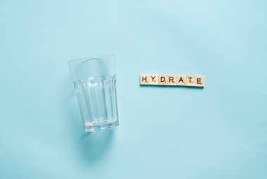 Minimalist flat lay of a drinking glass and letter tiles spelling 'HYDRATE' against a blue background.